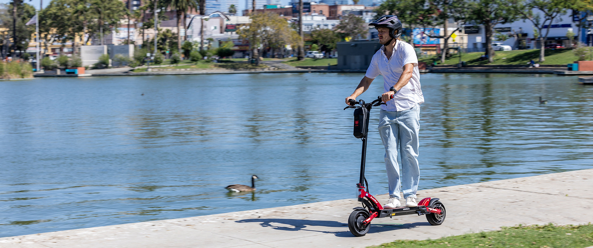 An adult male in casual attire and a helmet rides a red Kaabo Mantis 10 Lite electric scooter along a lakeside path. In the background, there are city buildings, trees, and a lake with ducks.