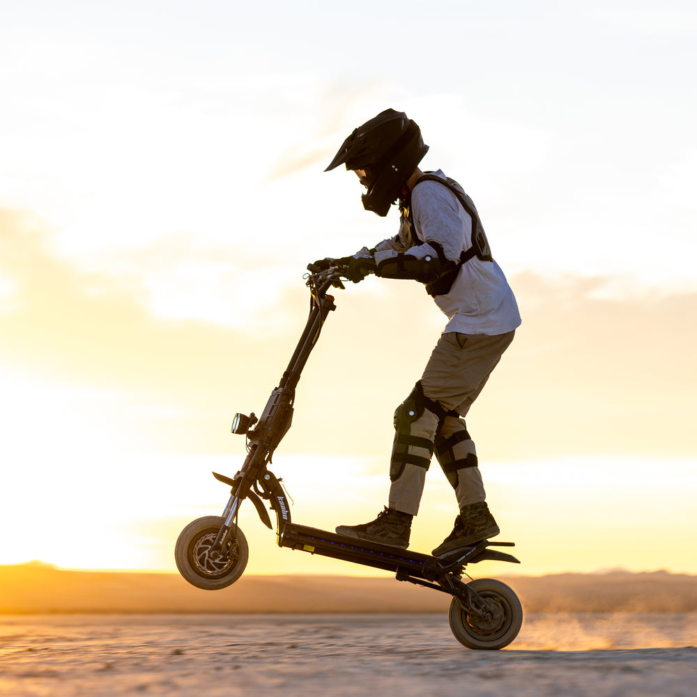 A person wearing a helmet and protective gear rides a black Kaboo electric scooter on a flat desert surface at sunset, with the scooter performing a wheelie.