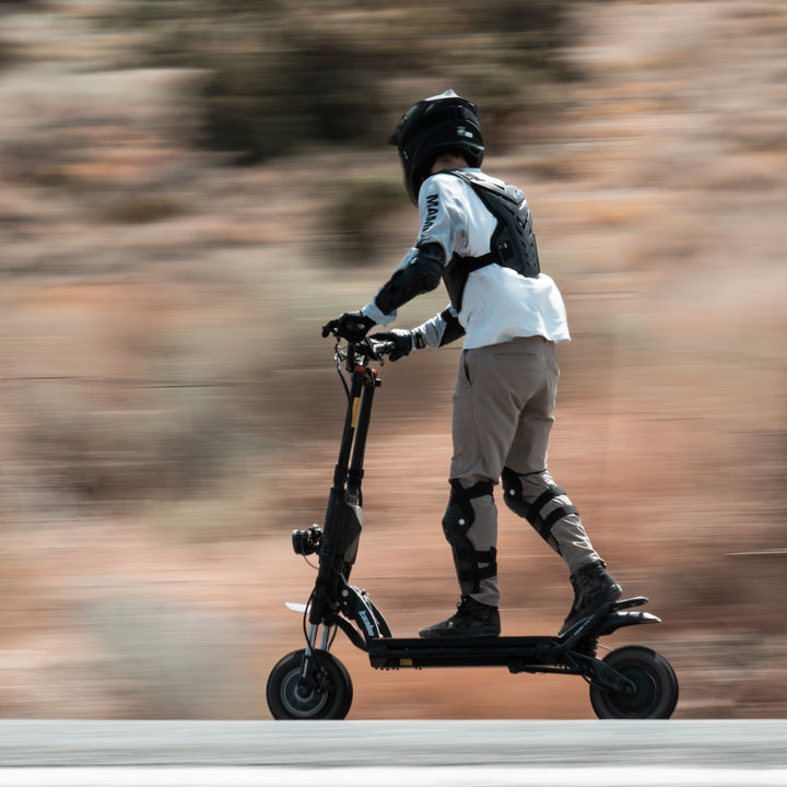 A person wearing a black helmet and protective gear rides a black electric scooter on a road with a blurred outdoor background.