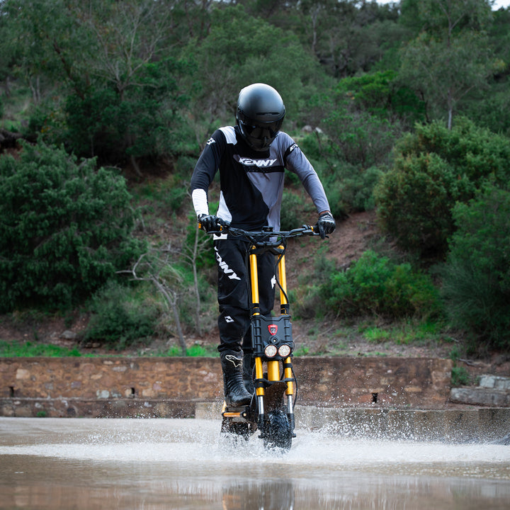 A person wearing a black and gray riding suit, helmet, and gloves operates a yellow and black electric dirt bike while splashing through shallow water outdoors, with trees and shrubs in the background.