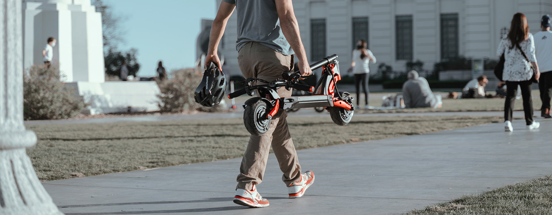 Commuter carrying a folded Kaabo Mantis 8 electric scooter across a city park, showcasing the portable design against an urban backdrop.