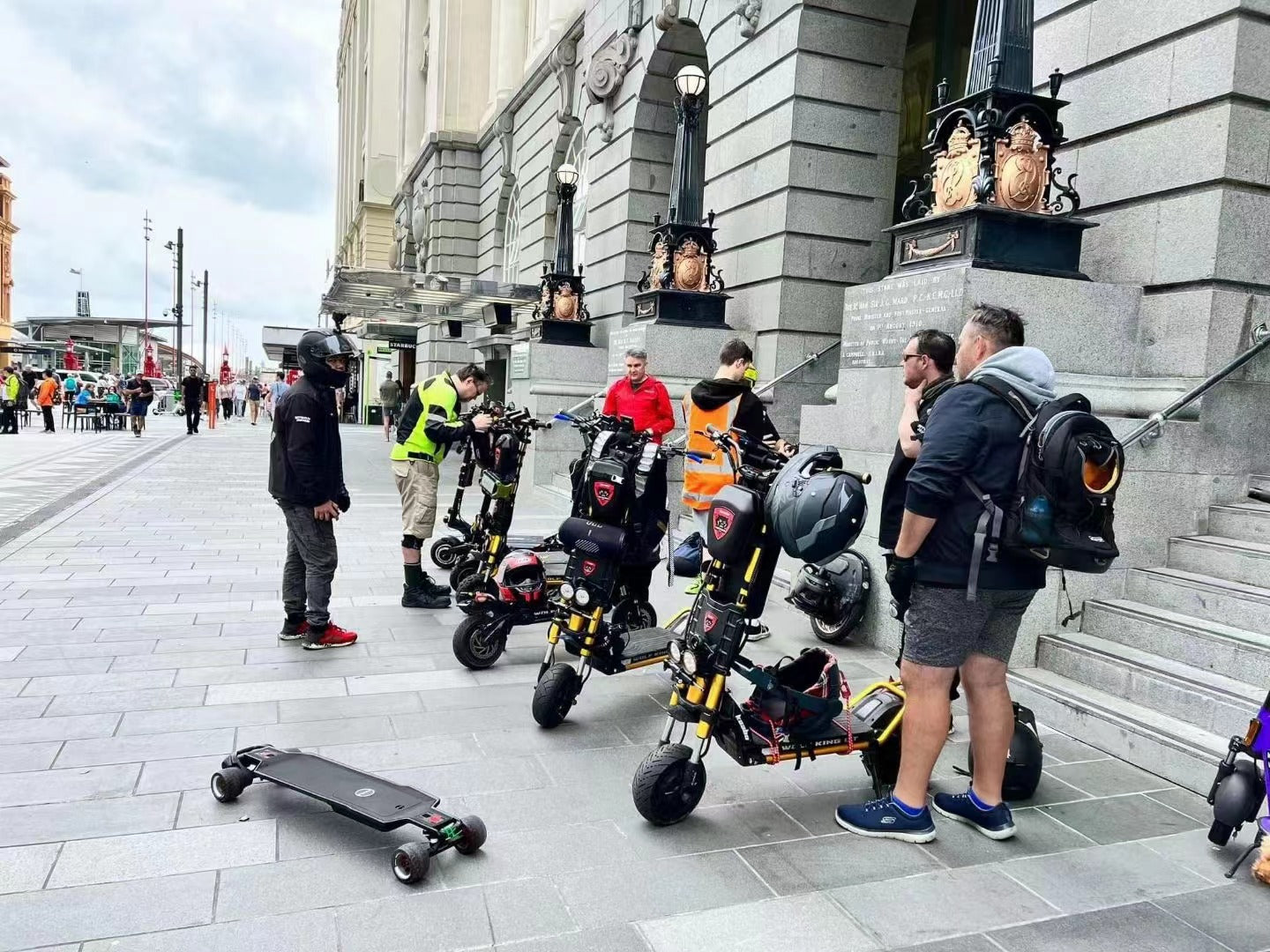 A group of individuals with electric scooters and skateboards congregating on a city sidewalk, engaged in conversation and examining their vehicles.