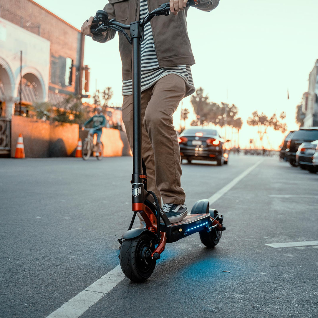 Person riding a Kaabo Mantis 8 electric scooter on a city street at dusk, showcasing the scooter's sleek design and urban functionality.