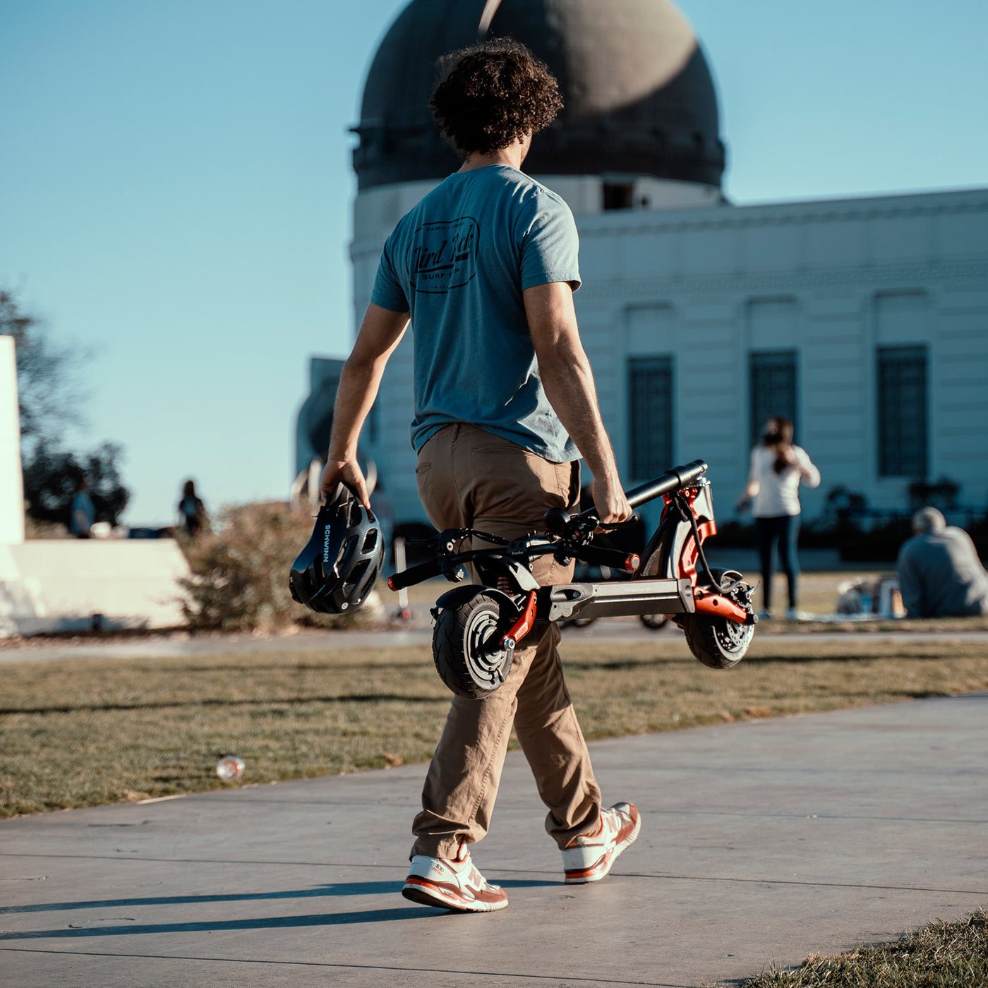 Man carrying a folded Kaabo Mantis 8 electric scooter in an urban park setting, illustrating the scooter's portability and compact design.