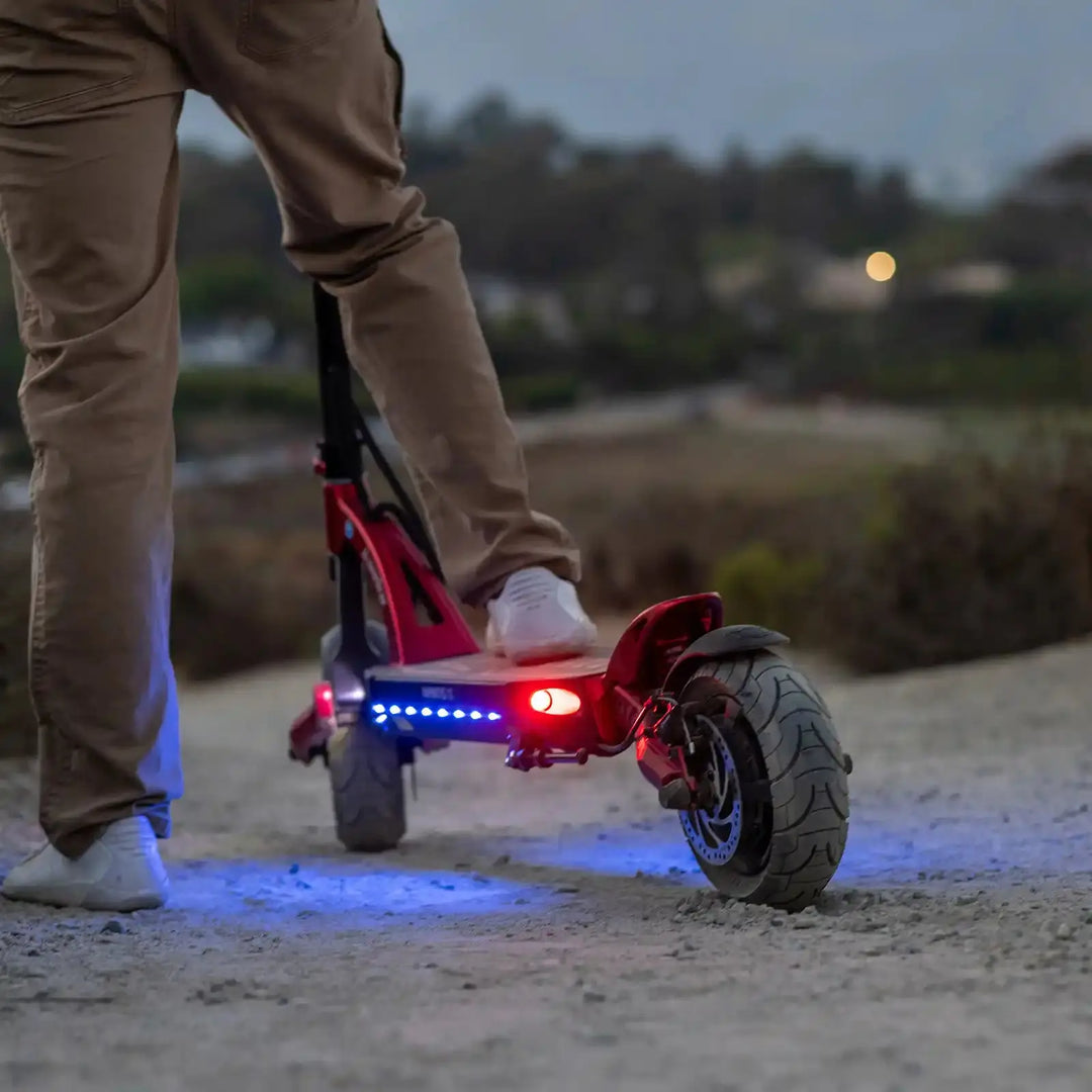 Person riding a red kaabo mantis 10 electric scooter with lights on a path at dusk.