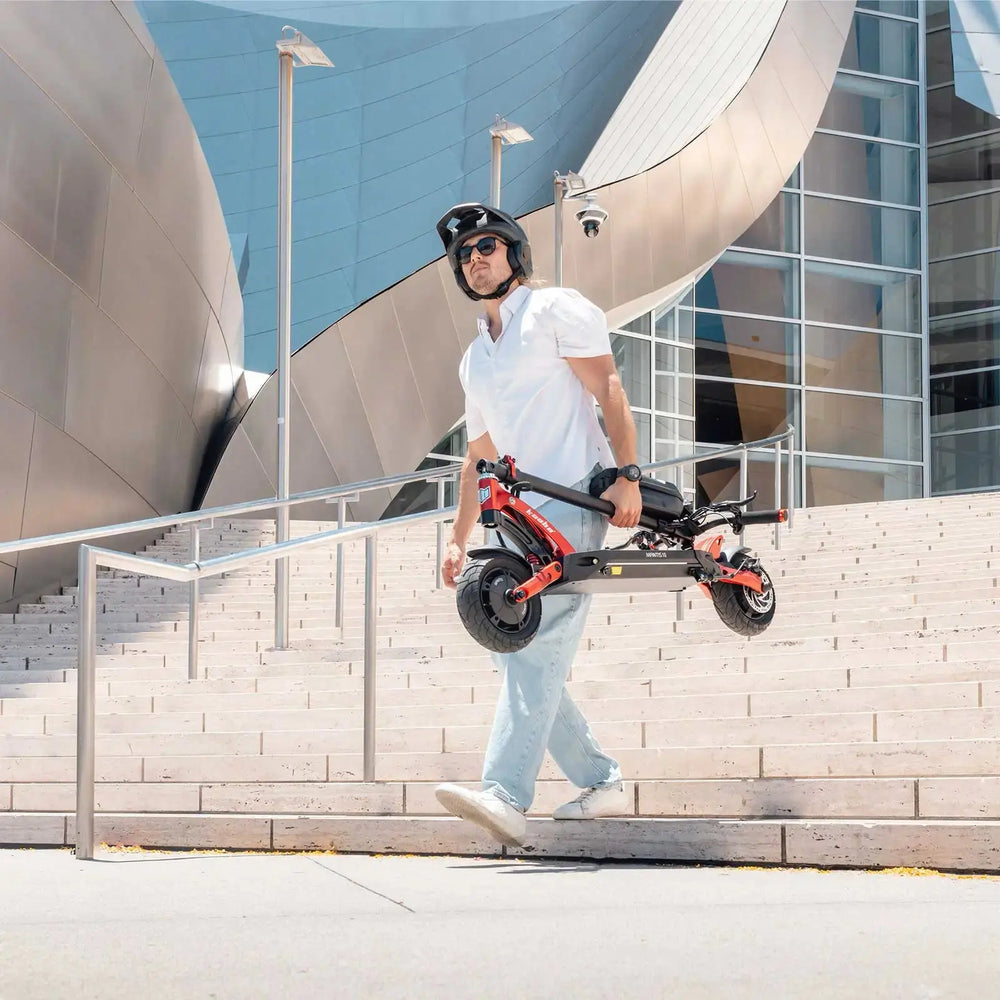 Man carrying Kaabo USA Mantis 10 Lite Electric Scooter in front of modern urban architectureA man wearing a helmet rides a Kaabo Mantis 10 Lite electric scooter along a lakeside path, with palm trees and buildings in the background.