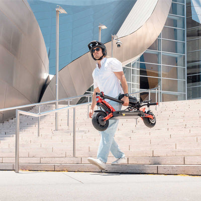 Man carrying Kaabo USA Mantis 10 Lite Electric Scooter in front of modern urban architecture