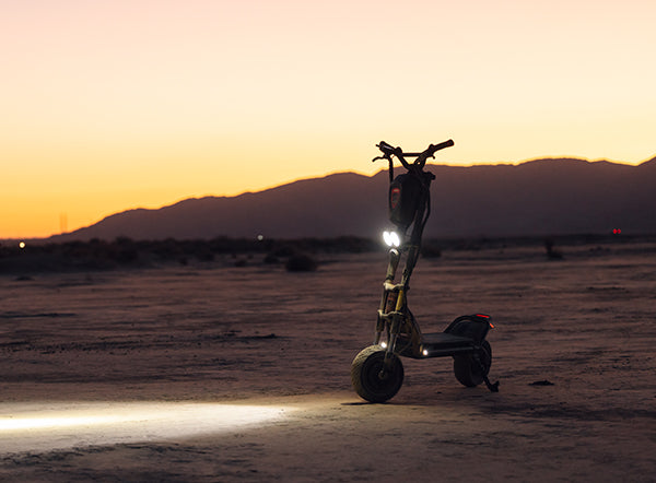 The headlight of a Kaabo Wolf Warrior 11 Pro+ electric scooter illuminating the ground at dusk with mountains silhouetted in the background.